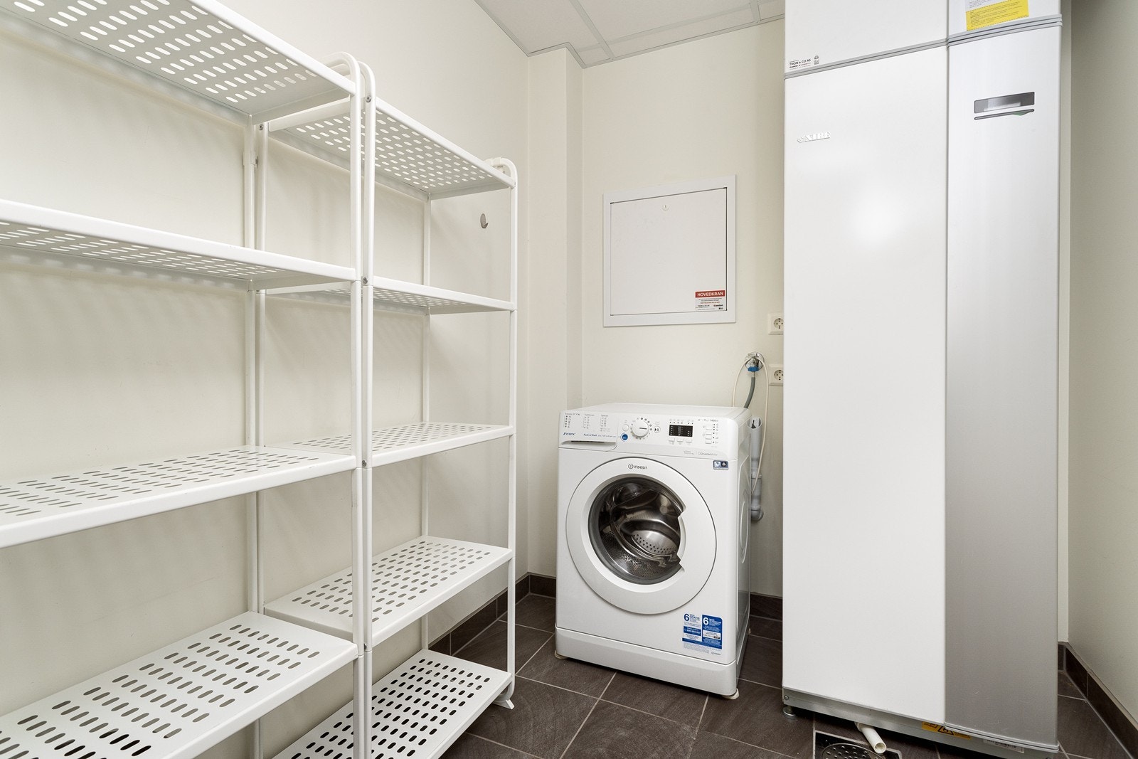 Praktisk vaskerom med flislagt gulv med opplegg for vaskemaskin. Rommet har hylleløsninger, perfekt for oppbevaring av sesongrelatert utstyr. Galleribilde