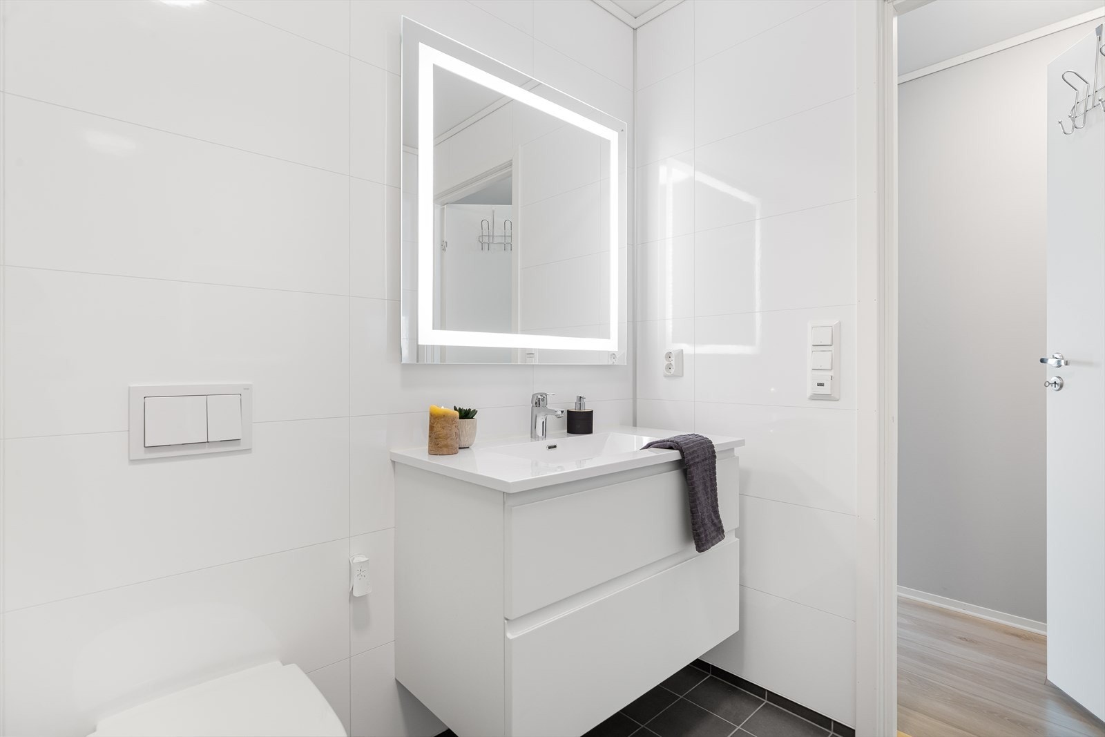 Lekkert bad med moderne servantskap og veggspeil med innebygd belysning. Mørke fliser på gulv med gulvvarme og hvite baderomsplater med flisprofil gir et stilrent og tidløst uttrykk. Galleribilde