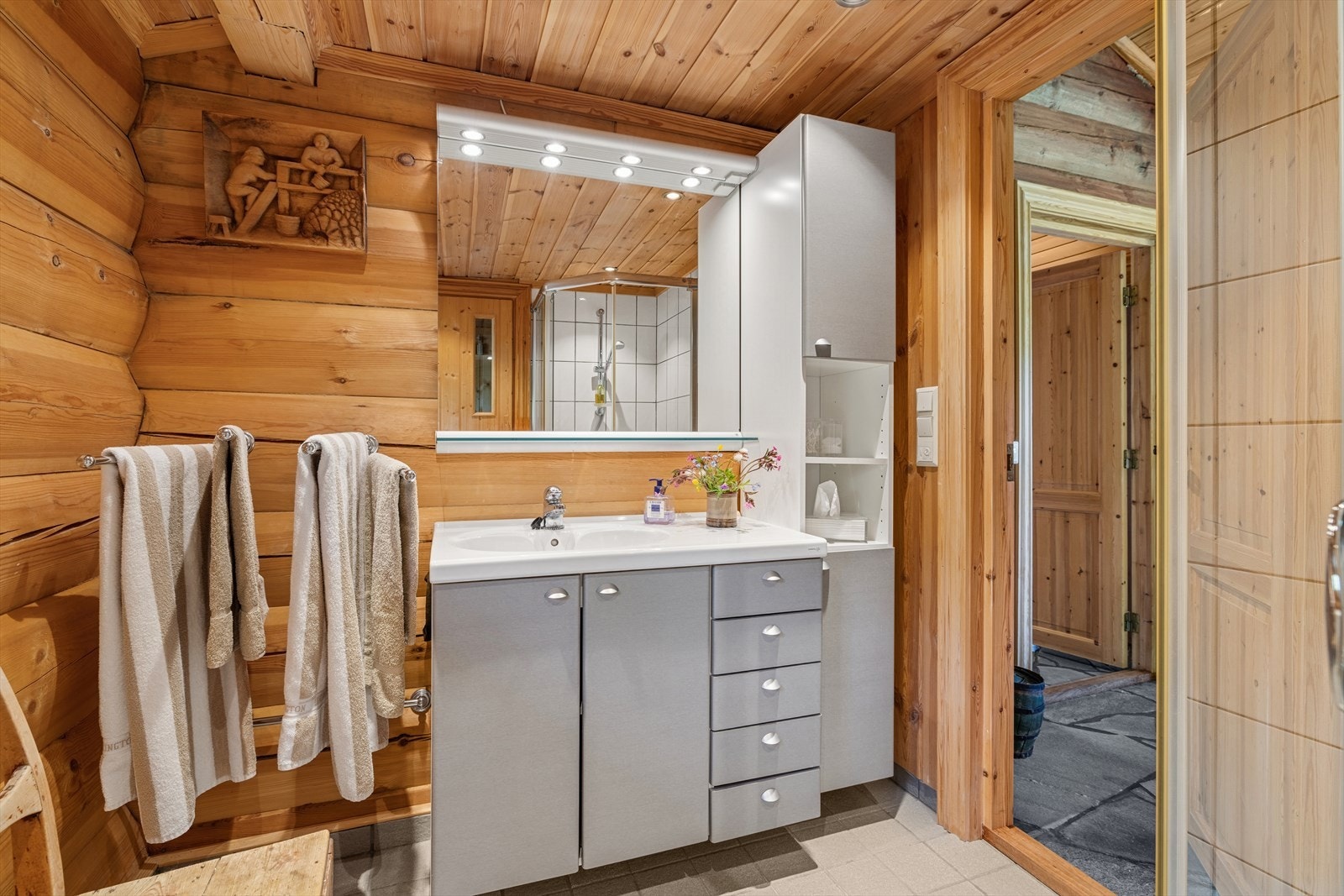 Badet i den nyere delen fremstår moderne og romslig, med flislagt gulv og behagelig gulvvarme. Tømmervegger kombinert med fliser i dusjsonen skaper en lun atmosfære. Galleribilde