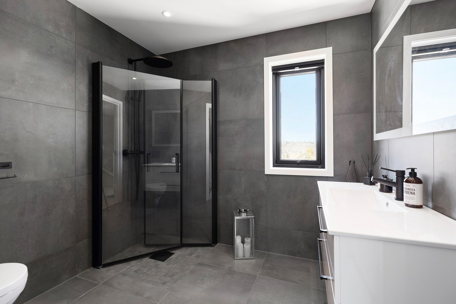 Badet er helfliset med mørke fliser som står i stil til resten av innredningen i boligen. Dusjhjørne, vegghengt toalett og servant i innredning. Galleribilde