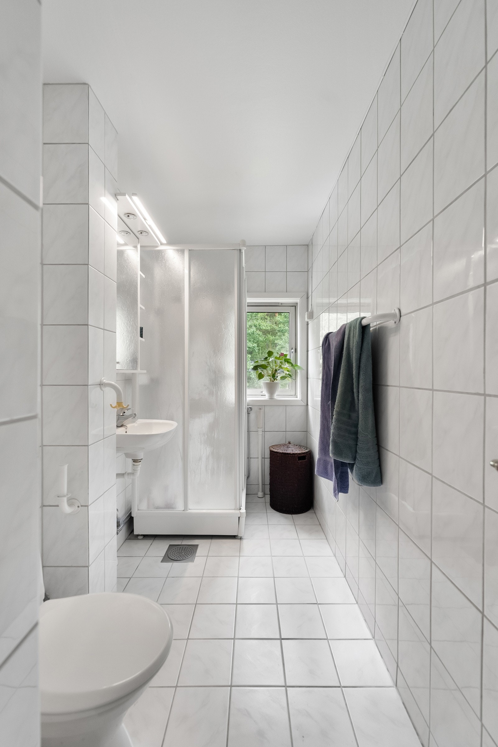 Opplegg for vaskemaskin er etablert, og badet har toalett og dusjkabinett. En enkel servant er plassert under et speilskap med tilhørende hyller og belysning. Galleribilde