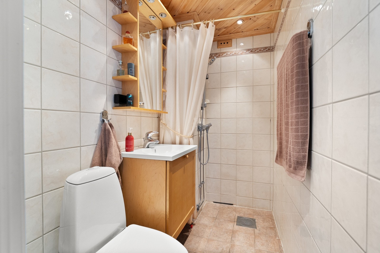 Badet i gangen er innredet med vegg- og gulvfliser i ulike sandtoner. Downlights er installert, og badet har toalett, varmekabler og et åpent dusjhjørne. Et servantskap i trestruktur er plassert under et matchende speilskap med tilhørende lys og hyller. Galleribilde