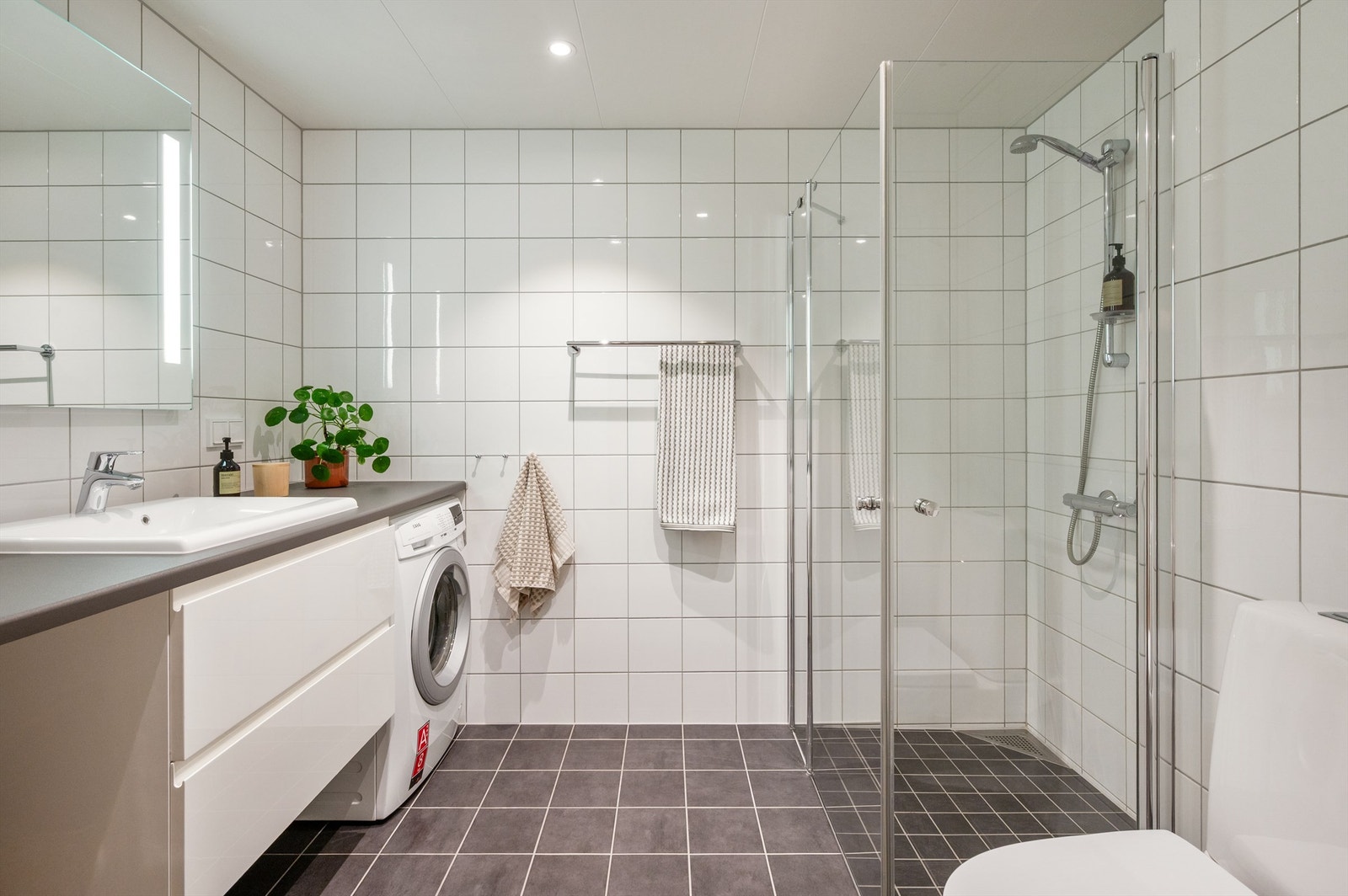 Badet er pent flislagt på gulv og vegger med varme i gulvet. Baderomsinnredningen består av vegghengt servantskap med servant. Videre har badet dusjhjørne med svingbare dører og klosett. Galleribilde