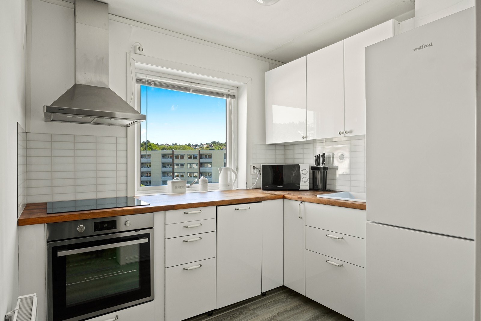 Kjøkkenet har en stilren og tidløs innredning med slette, hvite fronter fra 2016 som gir et moderne preg. Den solide benkeplaten i heltre tilfører varme og karakter, mens kitchenboard-plater med flisemønster på vegg gir et dekorativt og praktisk innslag. Galleribilde