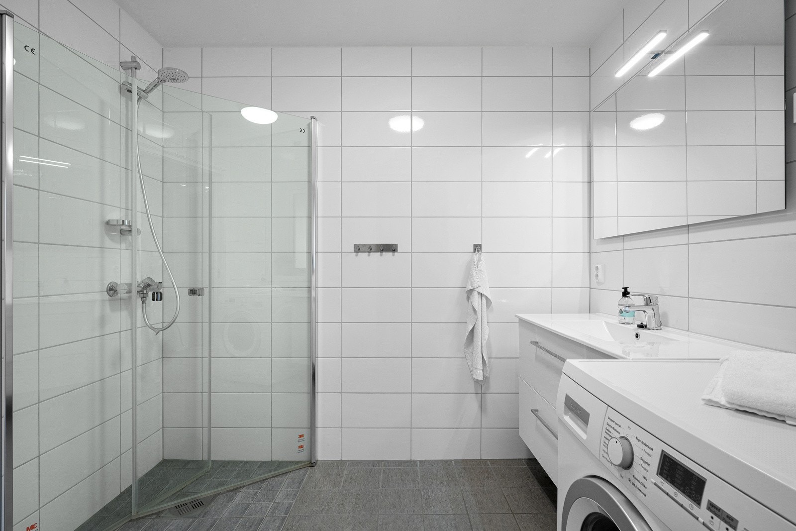 Badet har nedfelt servant med hylleseksjon, veggmontert toalett, dusjhjørne og opplegg for vaskemaskin. Galleribilde