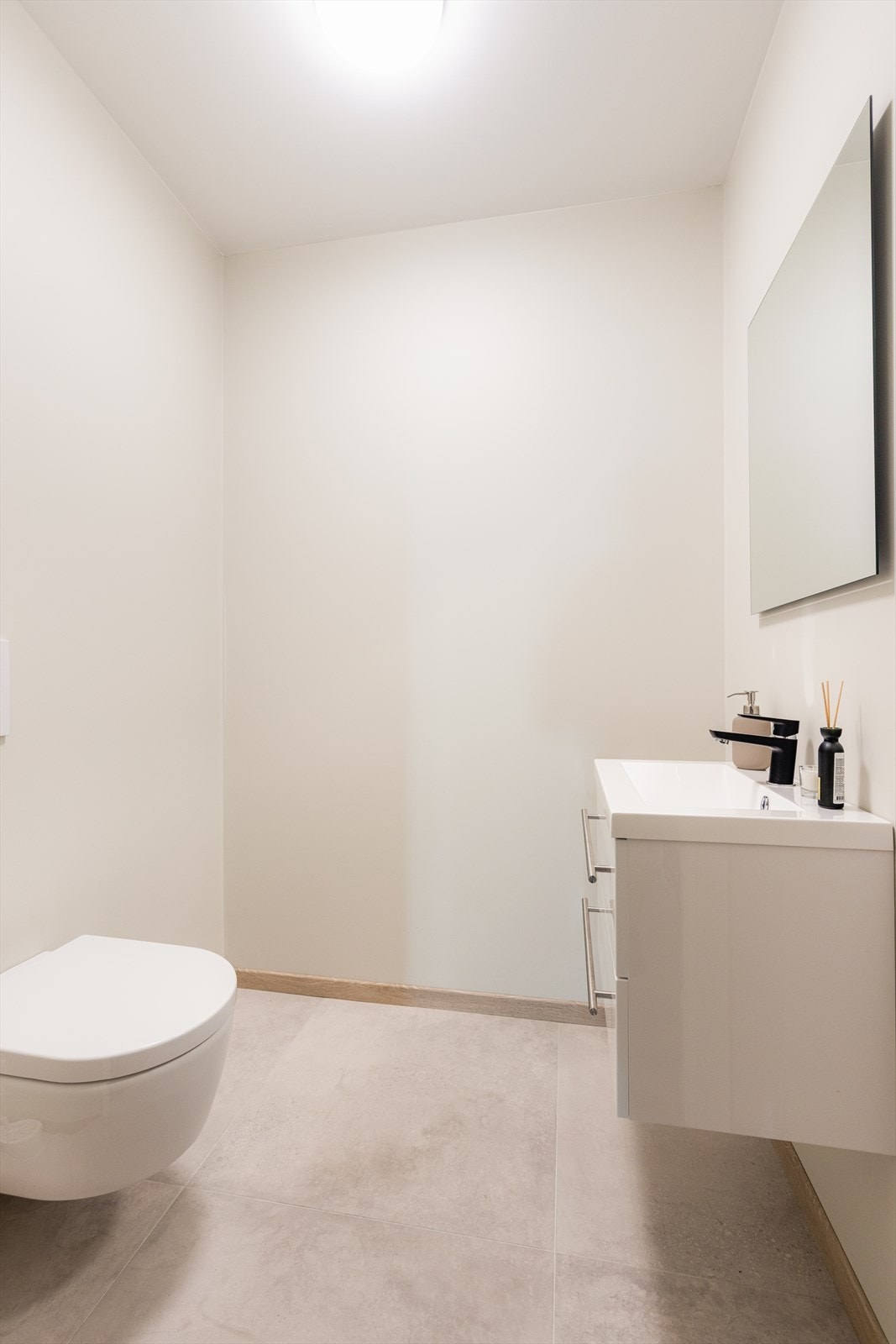 I enden av gangen er det et praktisk gjestetoalett med flis på gulv, veggmontert toalett og en tidløs baderomsinnredning med speilflate over vask. Galleribilde