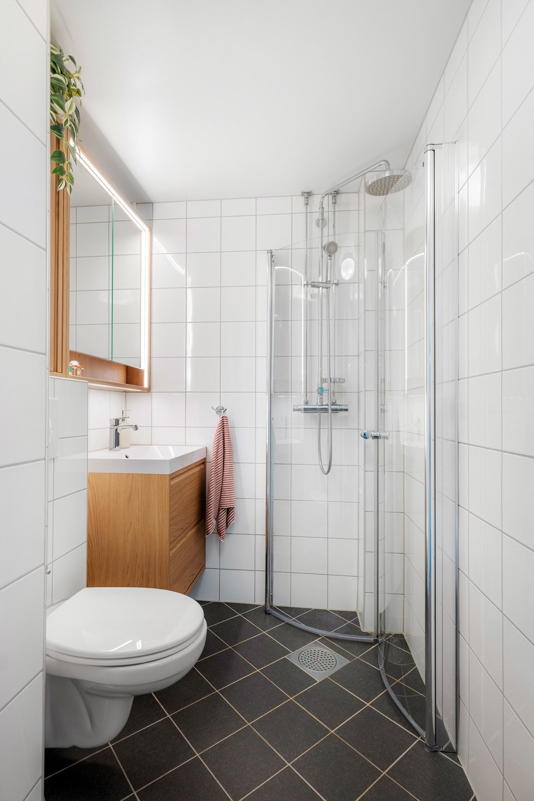 Baderom som inneholder: servantskap med glatte fronter, heldekkende servant, speilskap med lysarmatur over servant, veggmontert toalett, drenering i innkassing, dusjhjørne med svingdører i glass, sluk, dusjutstyr og termostatstyrt blandebatteri. Galleribilde