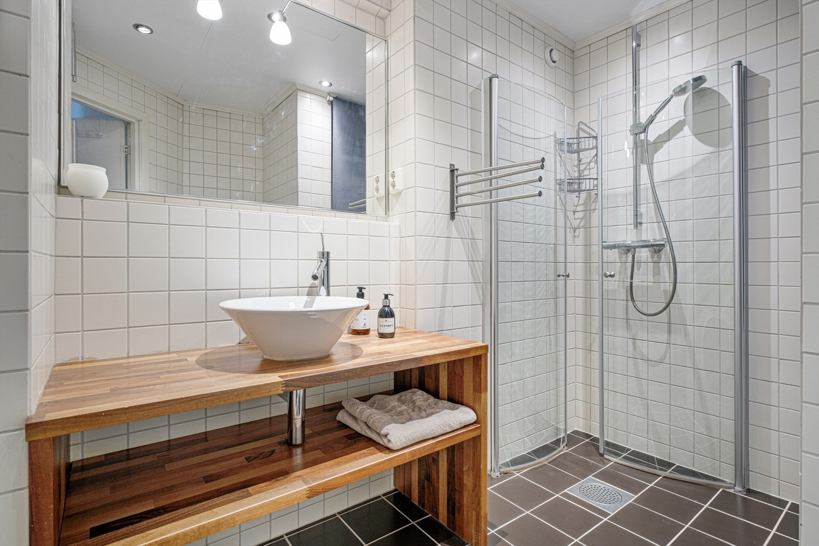 Pent flislagt bad som byr på praktiske innfellbare dusjdører av glass og dusjgarnityr på vegg, samt lekker innredning med servant, opplegg for vaskemaskin og veggmontert toalett. Galleribilde