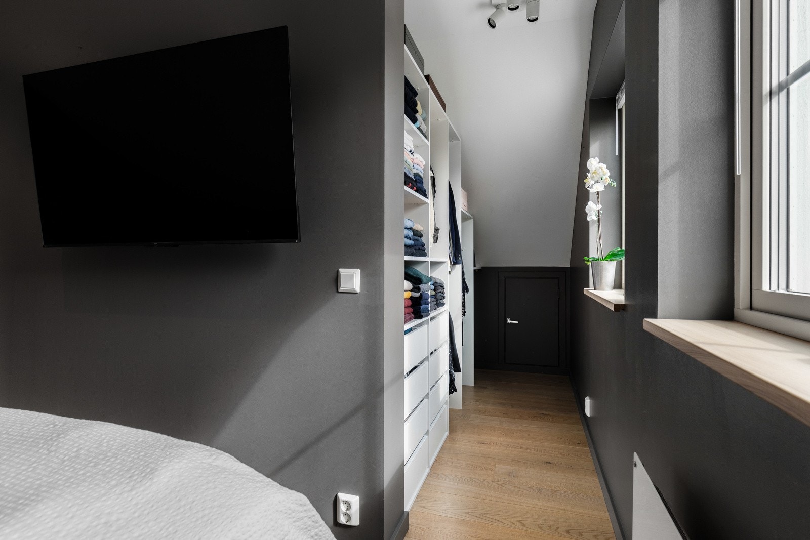 Rommet har mørke vegger, vindu, TV på vegg, og garderobe med hyller og skuffer. Lys parkett på gulvet. Galleribilde