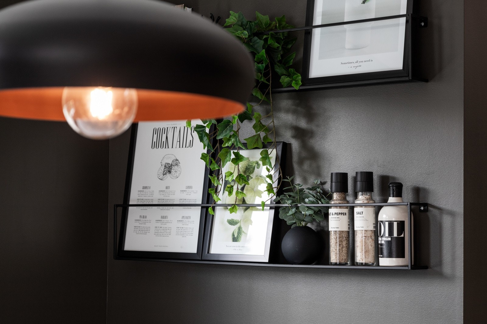 Svart hylle med plass til krydder og oljeflasker, dekorert med grønne planter og bilder, under en moderne lampe. Galleribilde