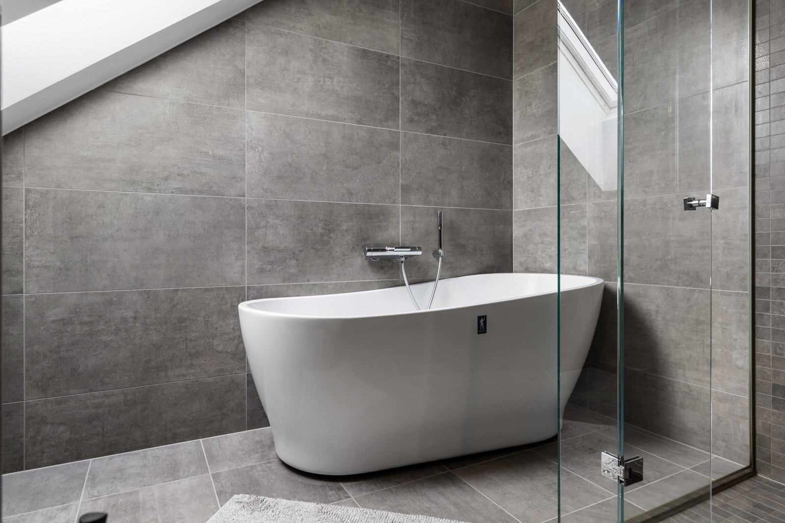 Moderne bad med frittstående badekar, grå fliser på vegger og gulv, samt dusjkabinett med glassdører. Galleribilde