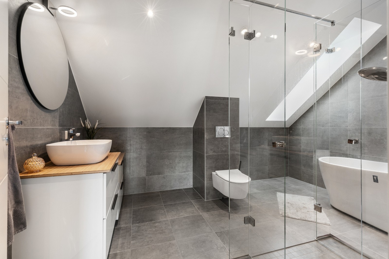 Moderne bad med grå fliser, dusjhjørne, frittstående badekar og servant på trebenk. Galleribilde