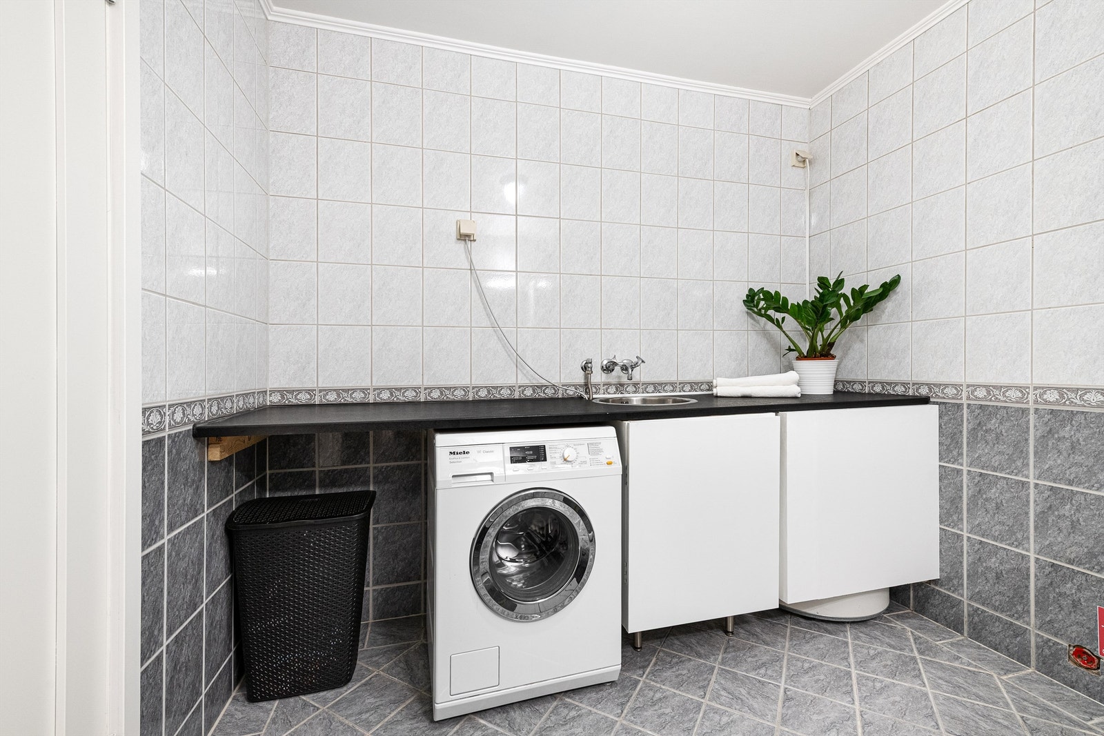 Praktisk med gulvstående skap og stor benkeplate over vaskemaskin med nedfelt utsalgsvask. Varmtvannsbereder er plassert i hjørne bak skap Galleribilde