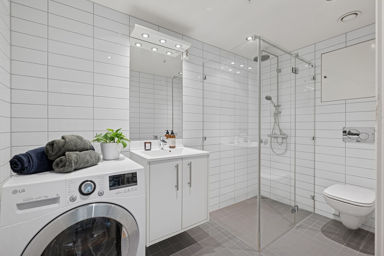Flislagt bad med vegghengt toalett, dusjsone og opplegg for vaskemaskin. Speil med integrert belysning og praktisk servantskap sørger for både komfort og oppbevaring. Galleribilde