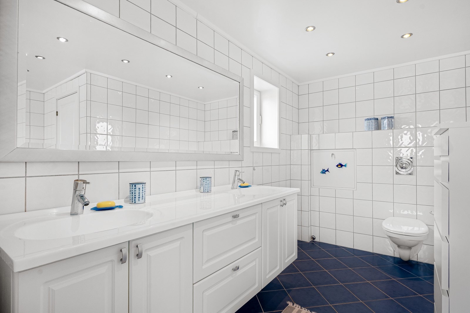 Badet har flislagte overflater i hvitt og blått, varmekabler og downlights. Det er god plass til håndklær og toalettartikler i innredning med to nedfelte servanter. Galleribilde