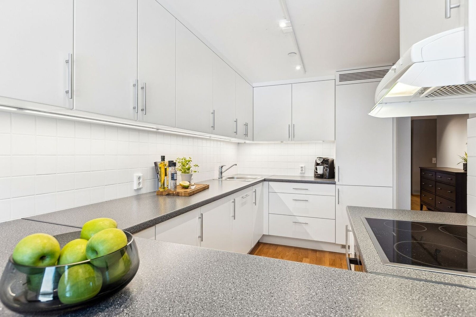 Her finner du et klassisk kjøkken med hvite, glatte fronter, belysning under overskap og stilige hvite fliser som gir et moderne uttrykk. Galleribilde