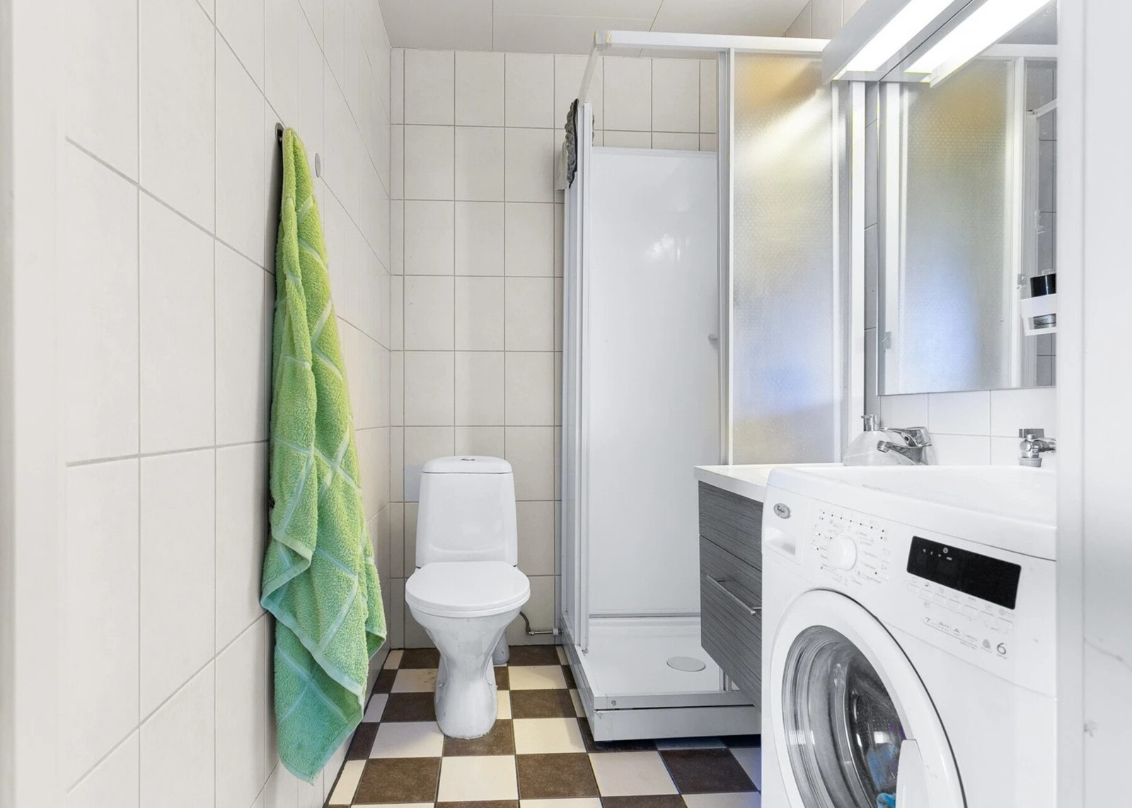 Bad med ukjent alder. Flislagt gulv og vegger. Malt himlingflate. Vegghengt servantskap med heldekkende servant og ett-greps armatur. Speil over servant med overlys. Dusjkabinett med dusjarmatur. Gulvmontert toalett. Opplegg for vaskemaskin. Galleribilde