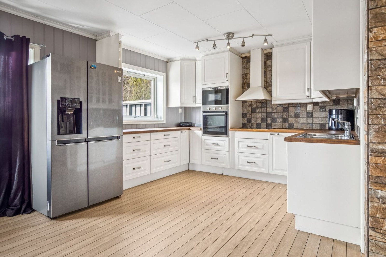 Lys kjøkkeninnredning med profilerte fronter, benkeplater i treverk, oppvaskkum med ett-greps blandebatteri og ventilator over kokesonen. Galleribilde