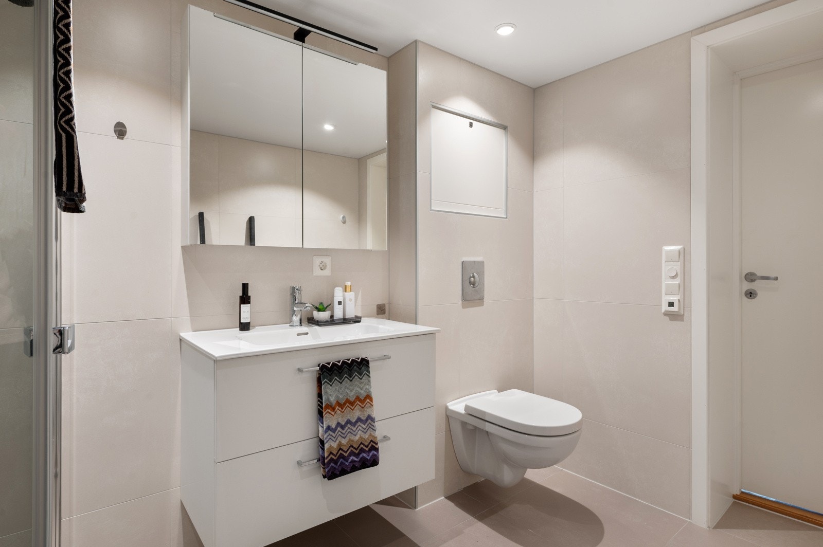 Badet har et moderne design og har varmekabler i gulv, innredning med servant og speilskap, veggfestet wc, dusj med dusjdører og opplegg til vaskemaskin Galleribilde