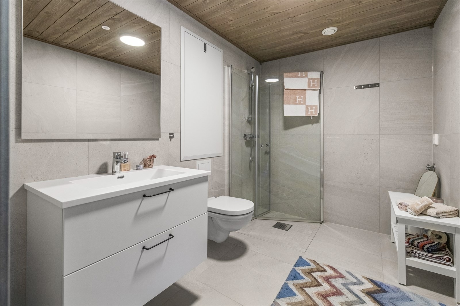 Badet er stilrent og moderne med store fliser på vegg- og gulvflater i en delikat beigefarge. Det er vannbåren gulvvarme, vegghengt toalett og opplegg for vaskemaskin. Innerst i rommet er det dusjhjørne med innfellbare glassdører og veggmontert hånddusj. Galleribilde