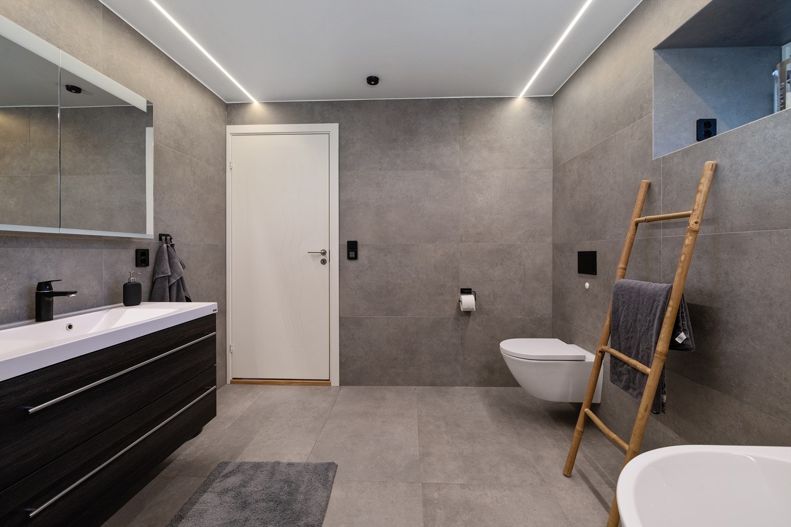 Badet er innredet med vegghengt toalett, badekar, dusjhjørne og servant på en moderne baderomsinnredning. Galleribilde