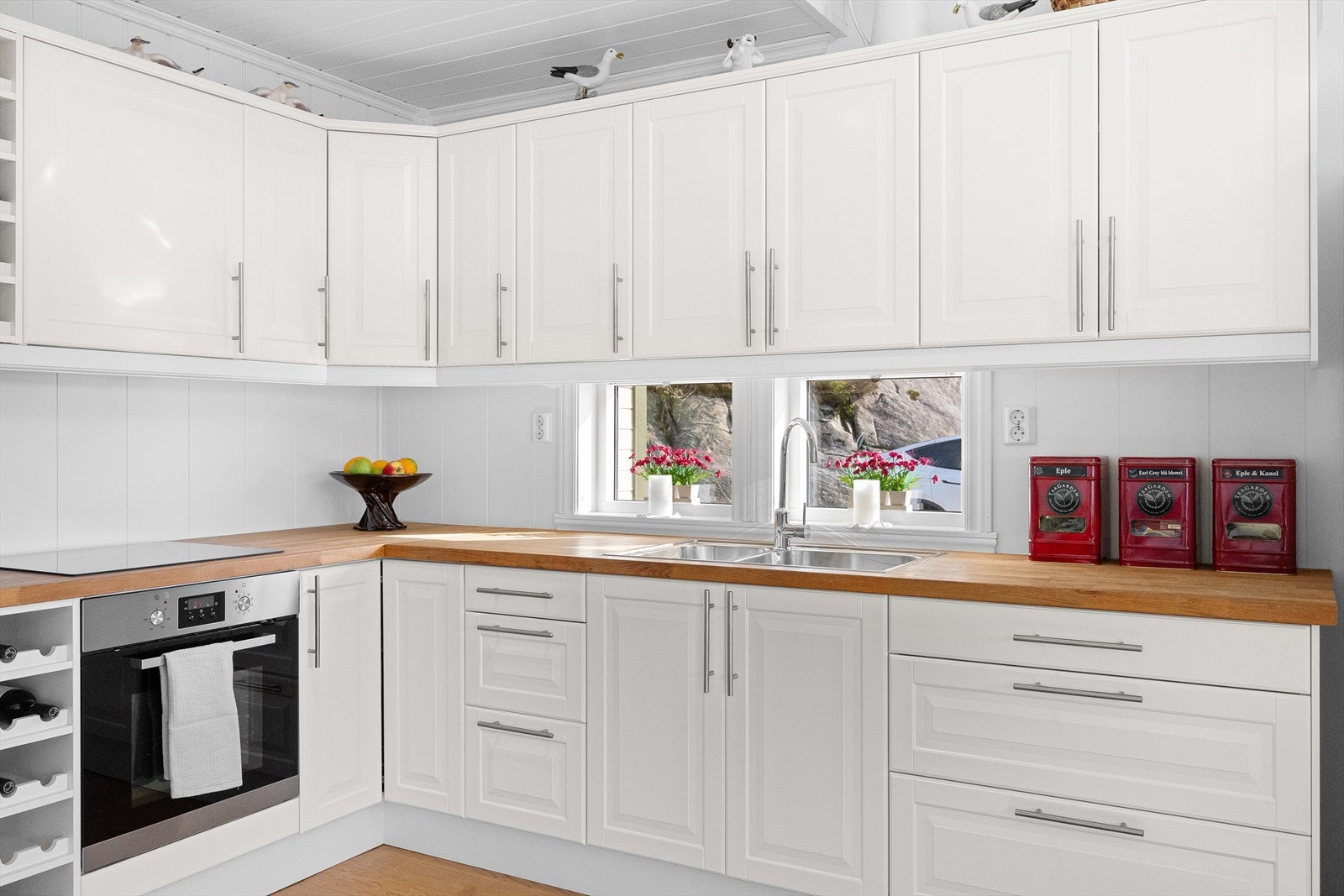 Lyst kjøkken med profilerte fronter og benkeplate med nedfelt stålvask og platetopp. Kjøkkenet er innredet med komfyr og kjøleskap. Galleribilde