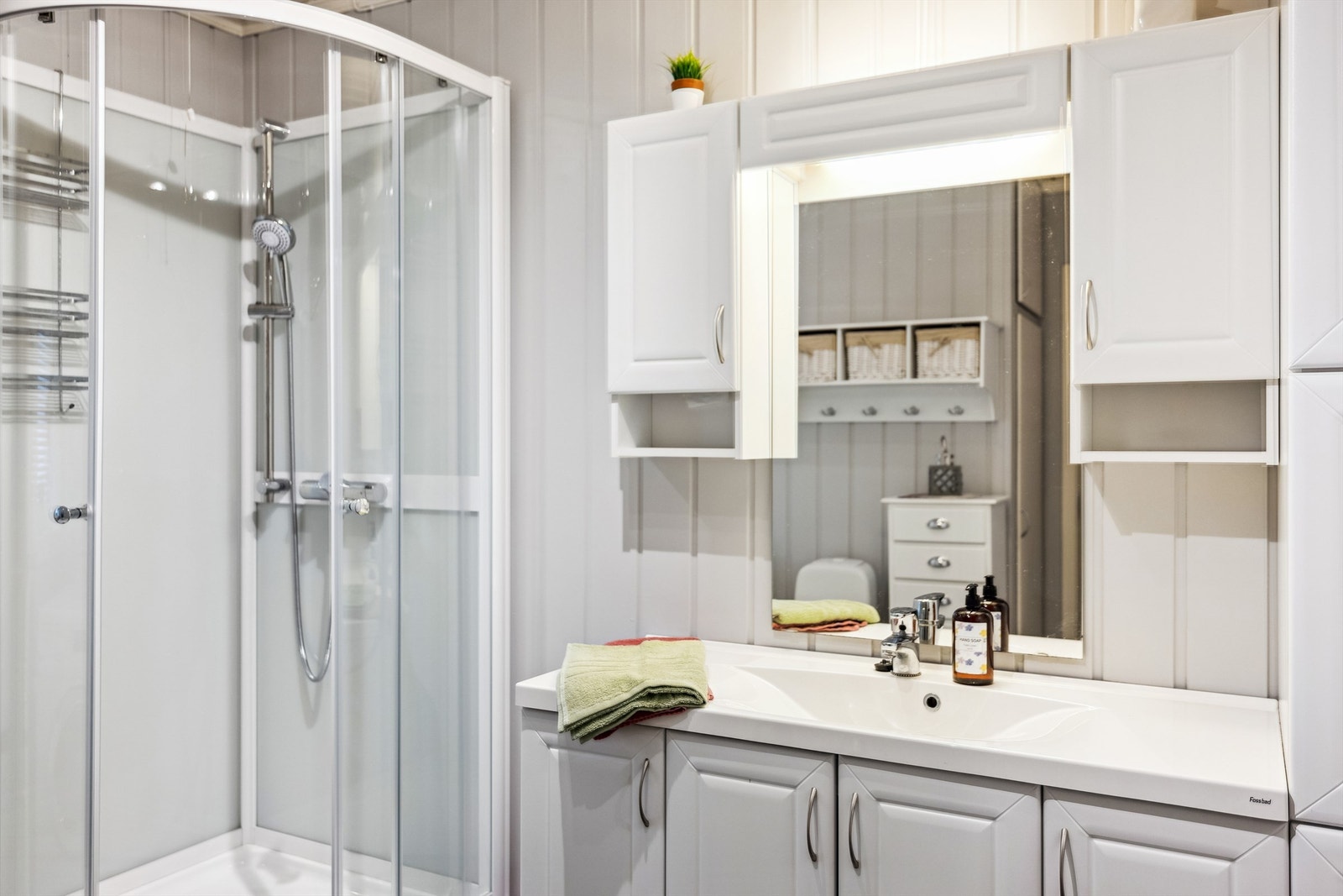 Badet er innredet med heldekkende servant i hvit skapinnredning, speil. vegghengte skap, dusjkabinett, gulvmontert toalett og opplegg for vaskemaskin. Galleribilde