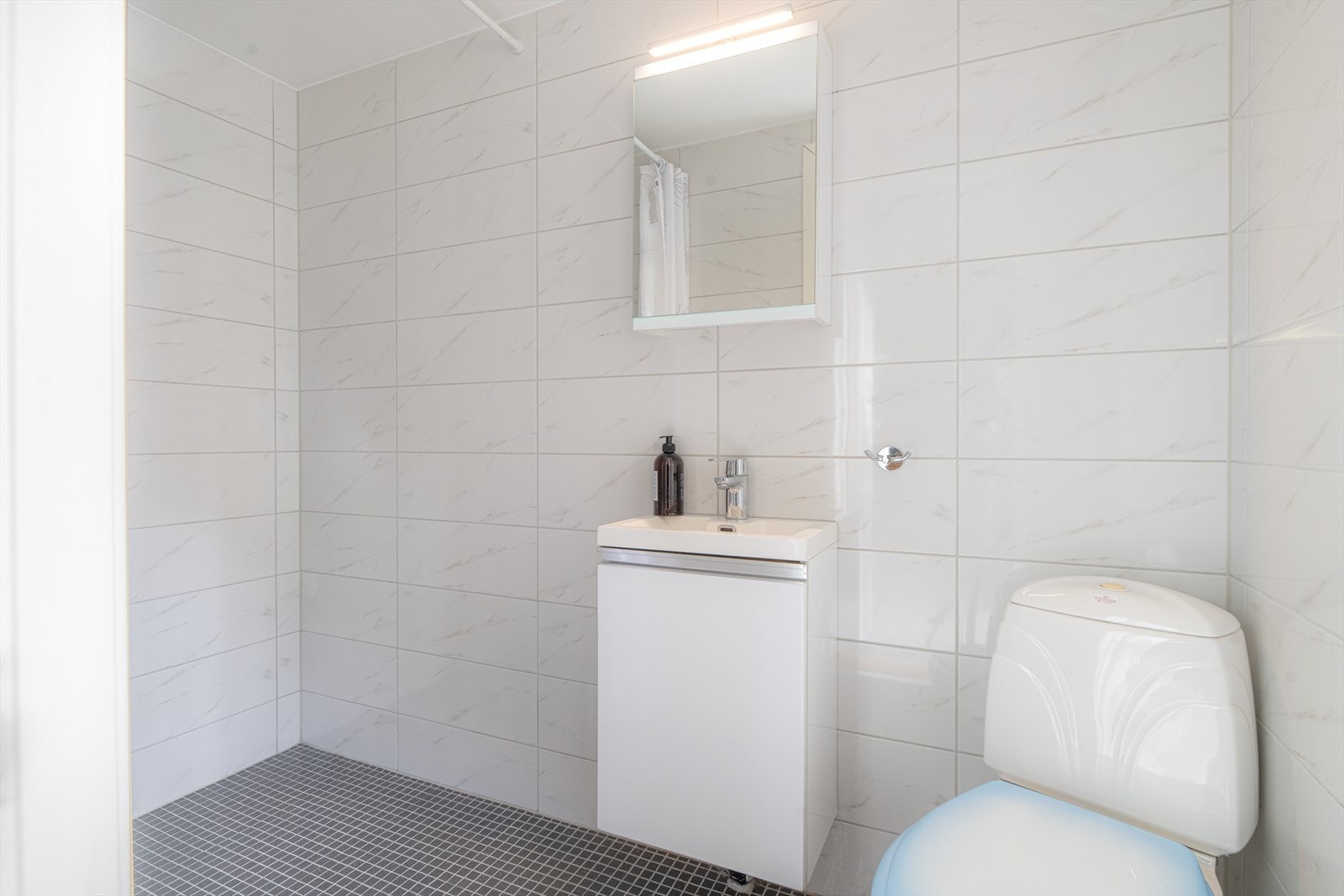 Badet er helfliset og har innredning med servant og speil med overlys, toalett og dusj på vegg. Galleribilde