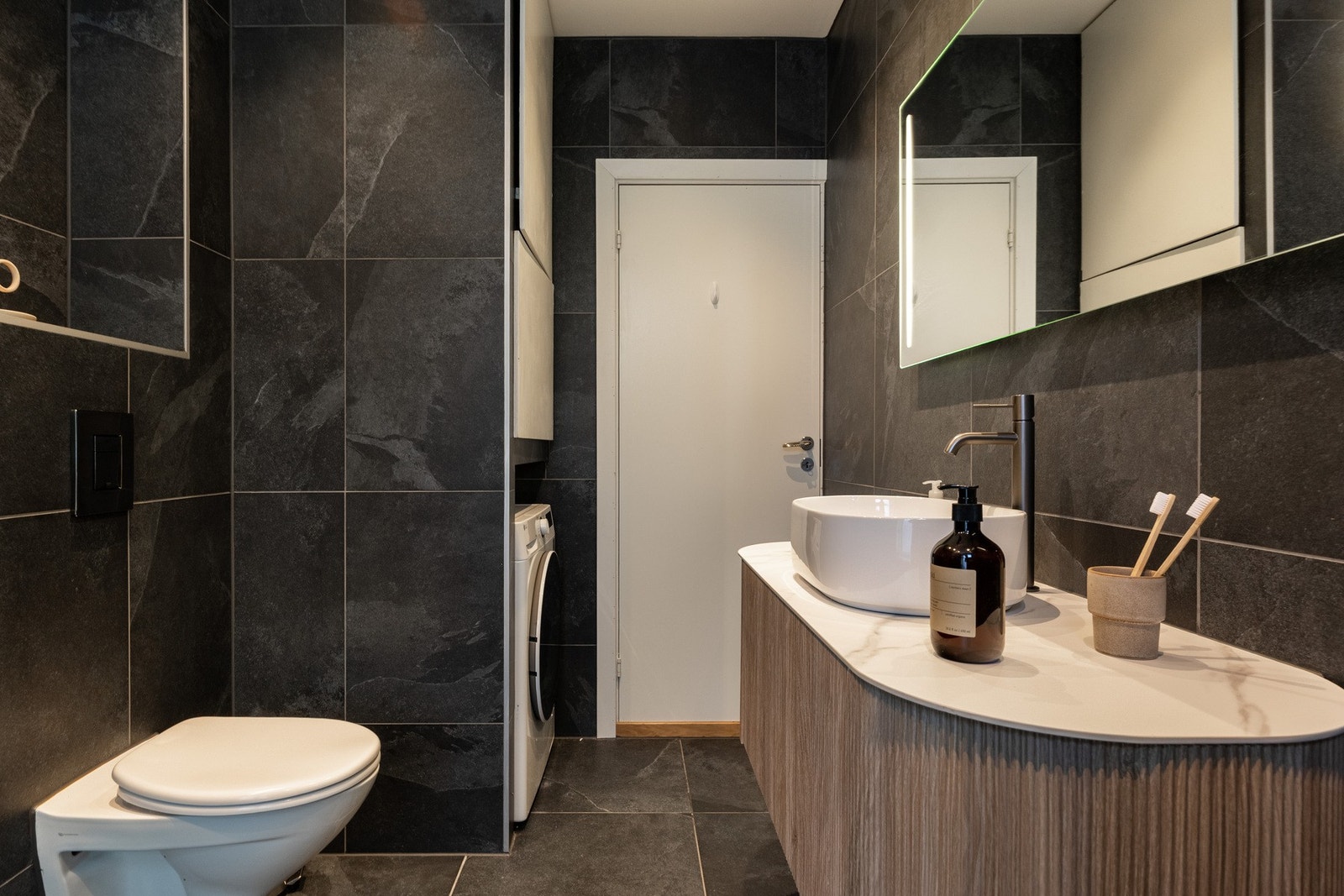 Badet er innredet med vegghengt servantinnredning med skuff, overliggende servant, speil på vegg med sidelys, dusjhjørne med glassvegg og regnfalldusj, samt vegghengt toalett med innebygd sisterne. Galleribilde