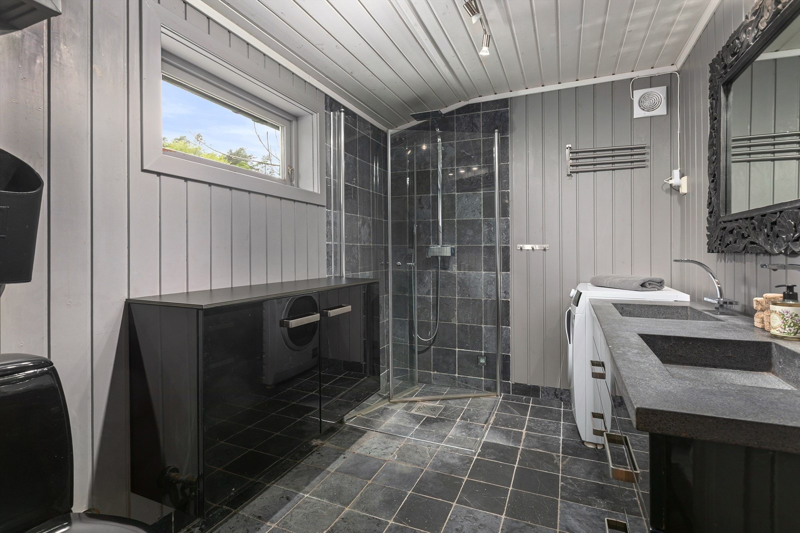 Badet med fliser på gulv og en kombinasjon av flis og malt panel på veggene. Det er opplegg for vaskemaskin og tørketrommel. Galleribilde