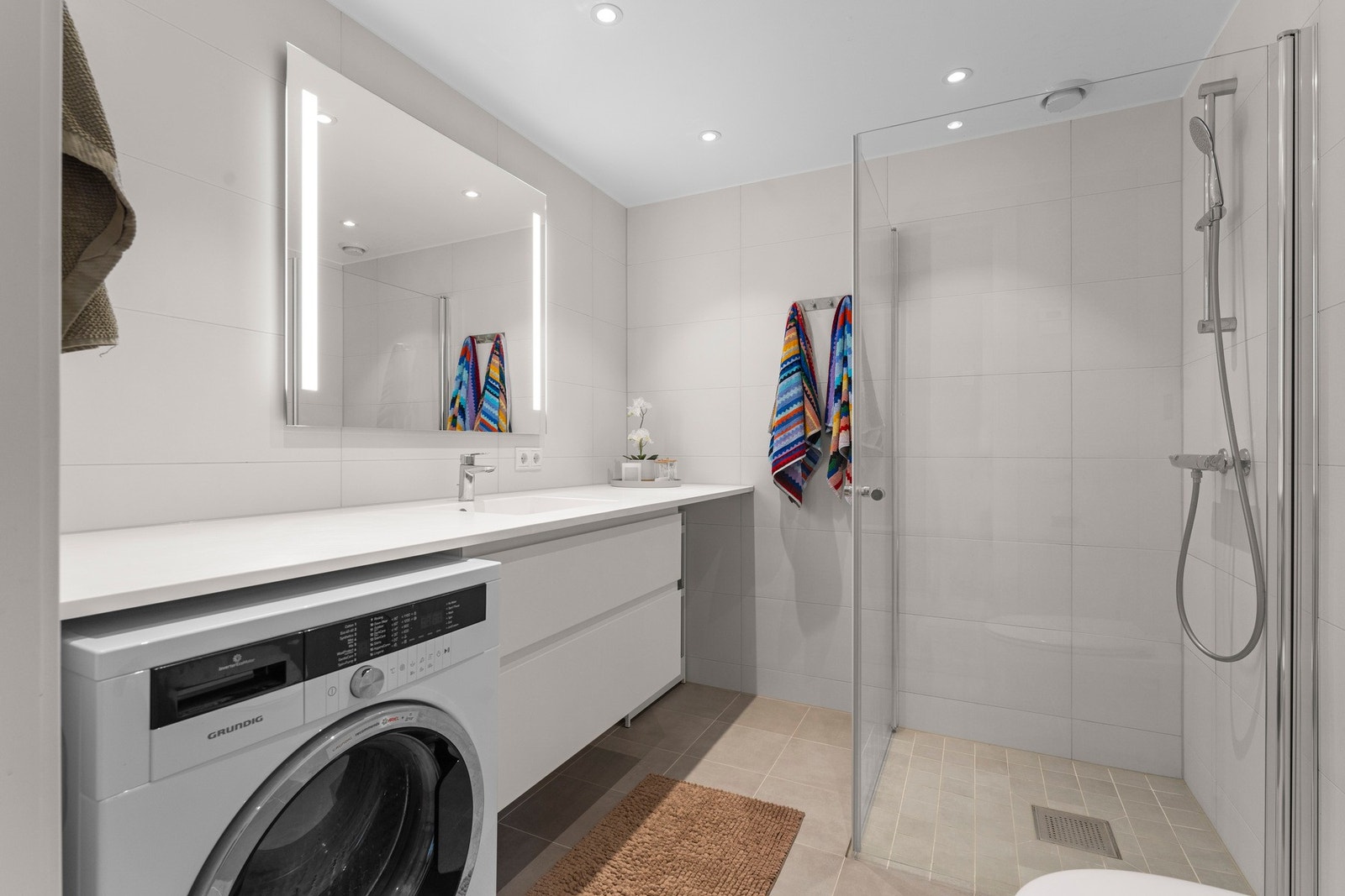 Badet har fliser på gulv og vegger, servant med benkeskap, veggmontert toalett og dusjhjørne med dører i herdet glass. Galleribilde