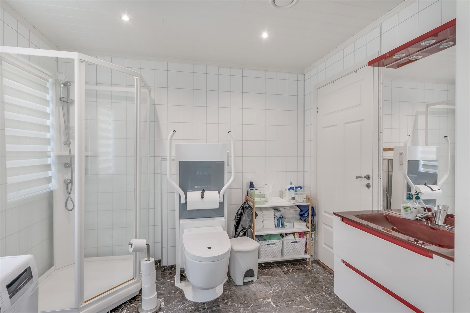 Badet har innredning med underskap, vask, høyskap og speil. Det er HC-toalett, dusjkabinett og opplegg for vaskemaskin. Inngang til teknisk rom. Galleribilde