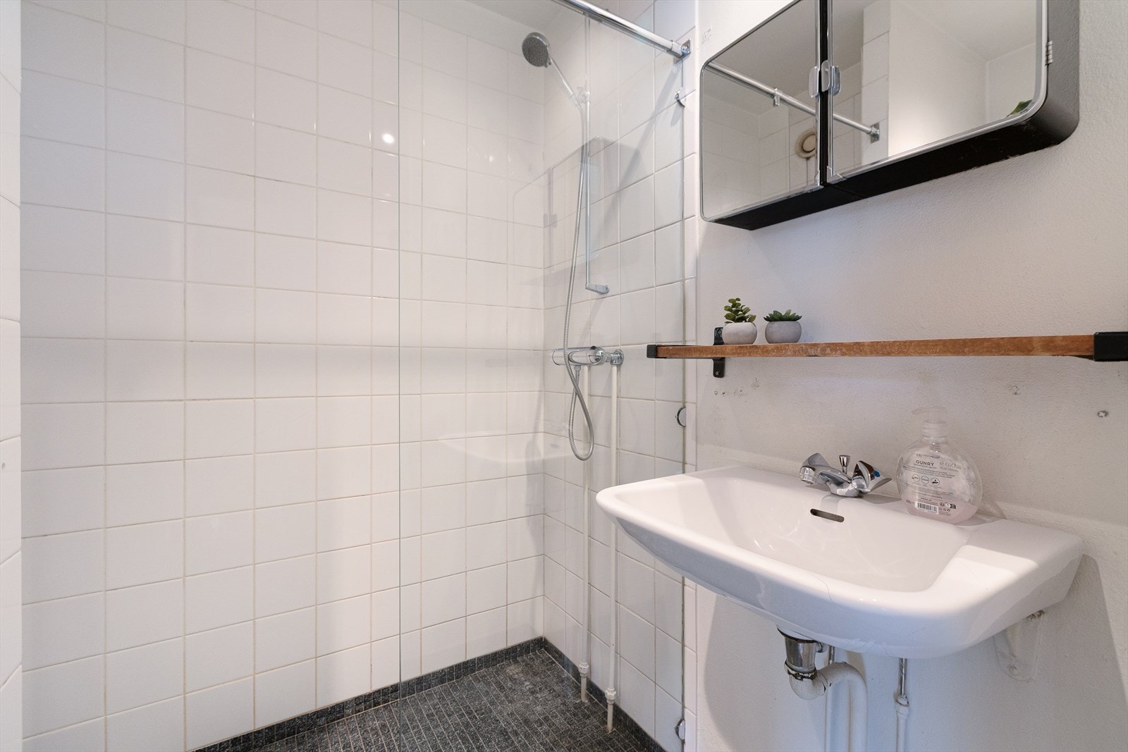 Flislagt baderom med romslig dusjsone, skillevegg av glass, toalett, servant og overhengte skap Galleribilde