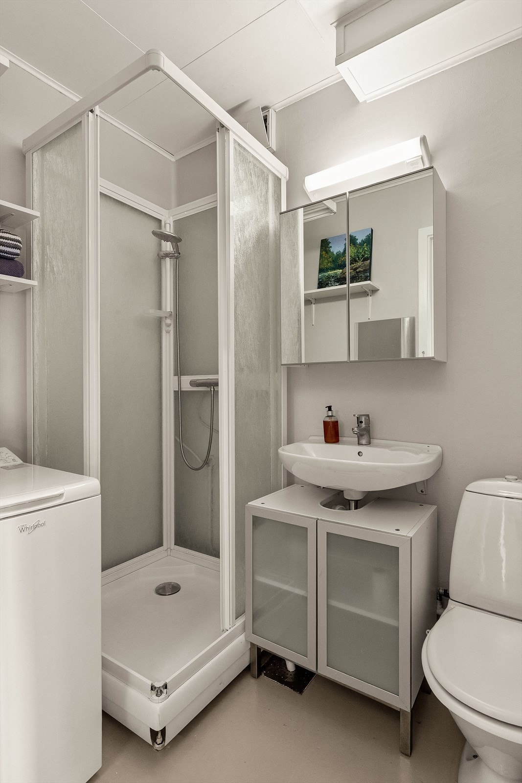 Lyst baderom med en innredning bestående av gulvmontert toalett, servant med underskap, speilskap, dusjkabinett og opplegg for vaskemaskin. Galleribilde