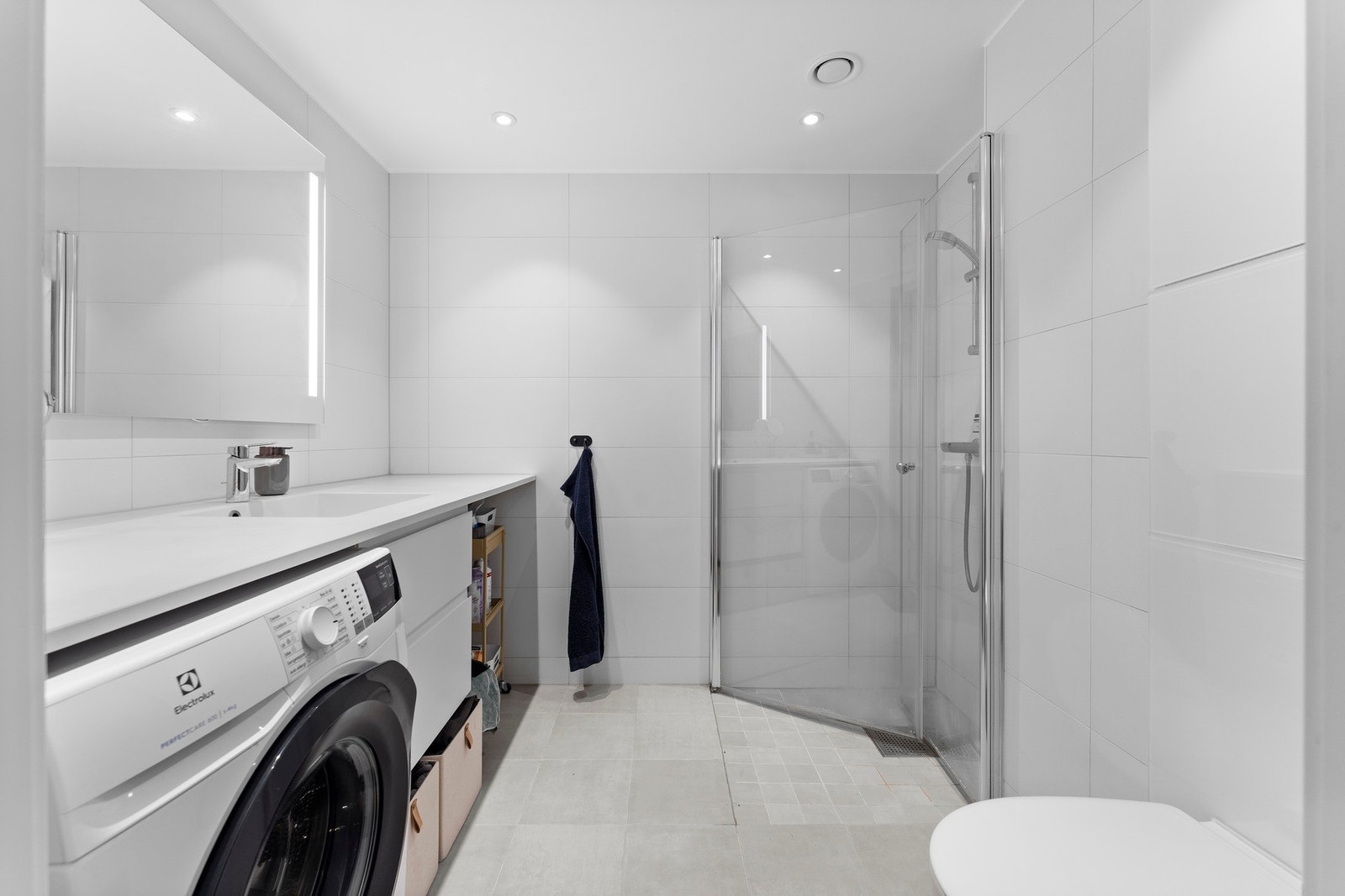 Badet har flislagte gulv og vegger som gir et rent og tidløst uttrykk. Her finner du servant med innredning og speil, vegghengt toalett, samt dusjhjørne med glassdører. Galleribilde