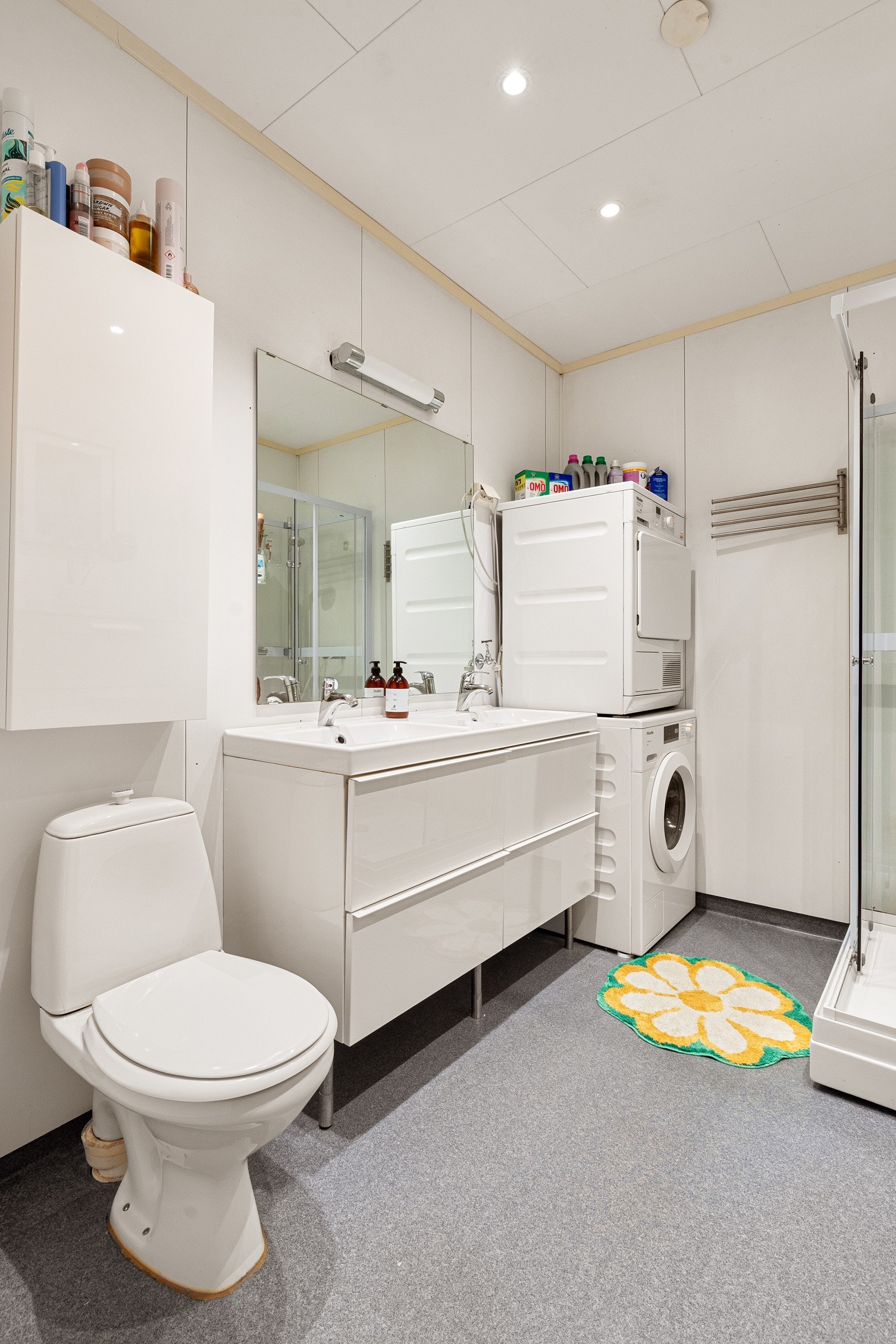 Moderne baderomsinnredning med skuffer og heldekkende dobbel servant, speil med belysning over. Baderommet er også utstyrt med dusjkabinett, toalett og opplegg for vaskemaskin. Galleribilde