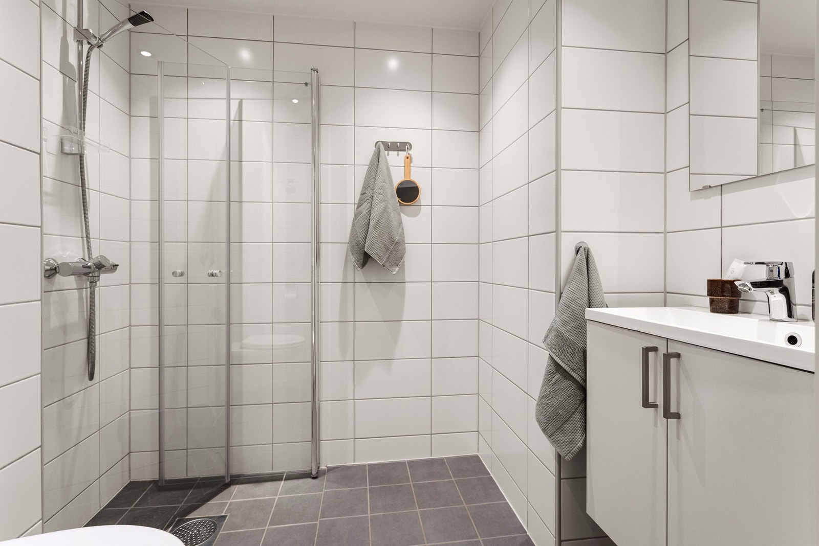 Badet er lyst og flislagt med gulvvarme. Innredningen består av servantskap, ovenpåliggende servant med ett-greps armatur, speil, belysning over speil, downlights, dusjsone og vegghengt toalett. Galleribilde