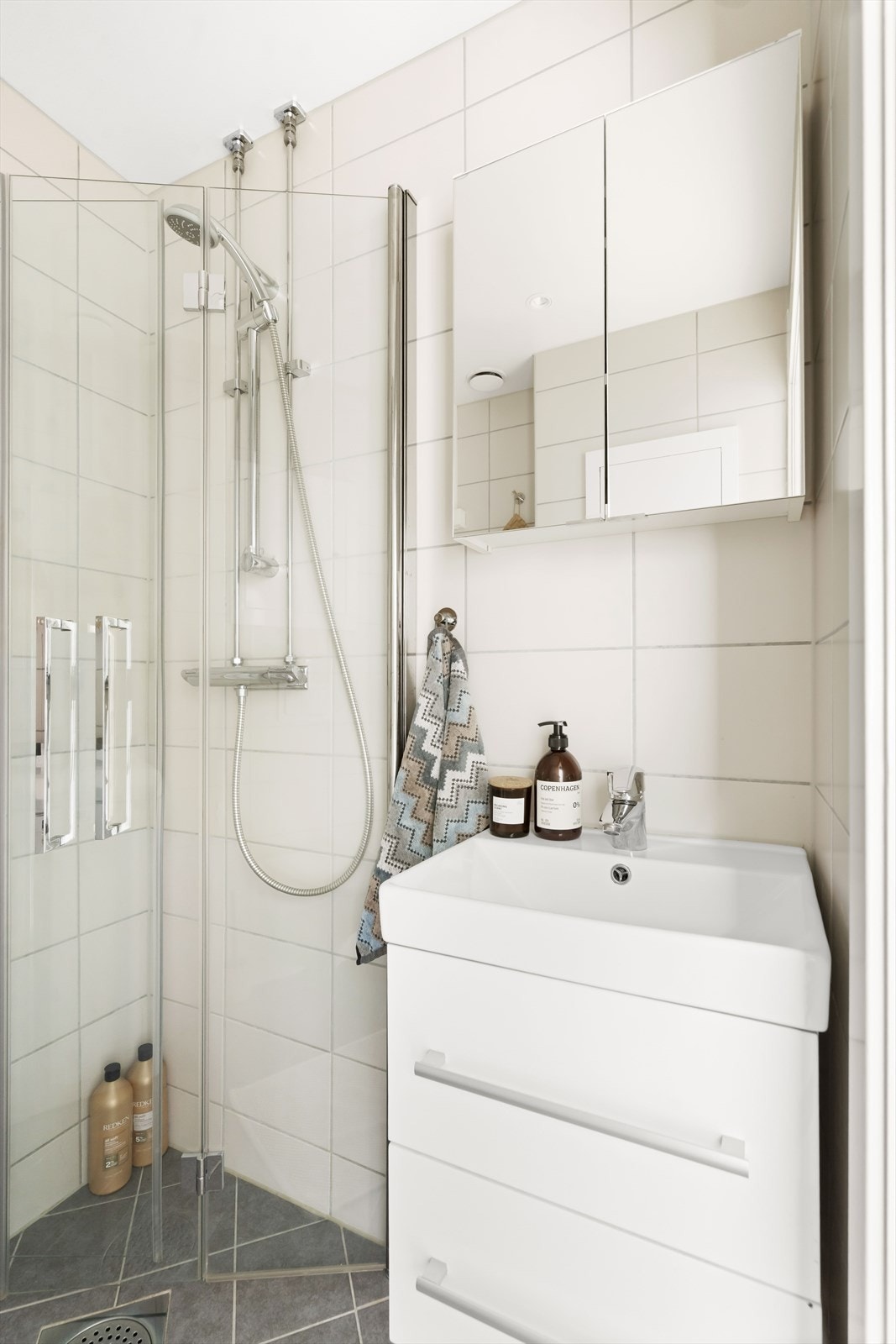 Badet har flislagte overflater, behagelig gulvvarme og innfelte downlights i himling. Badet er innredet med vegghengt toalett, dusjhjørne med svingbare glassdører og baderomsinnredning med skuffer, servant og speilskap. Galleribilde