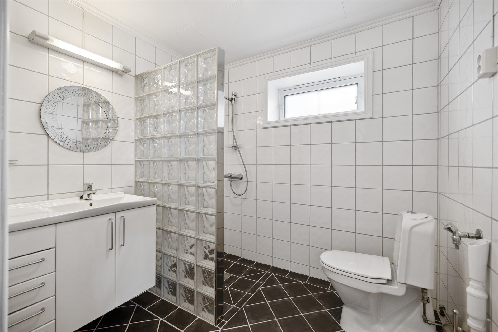 Badet i kjelleren er fra byggeåret og har fliser på gulv og vegger, samt himlingsplater i taket. Det er utstyrt med gulvmontert toalett, servant i hvit innredning og en dusjnisje med delevegg av glassbyggerstein. Galleribilde