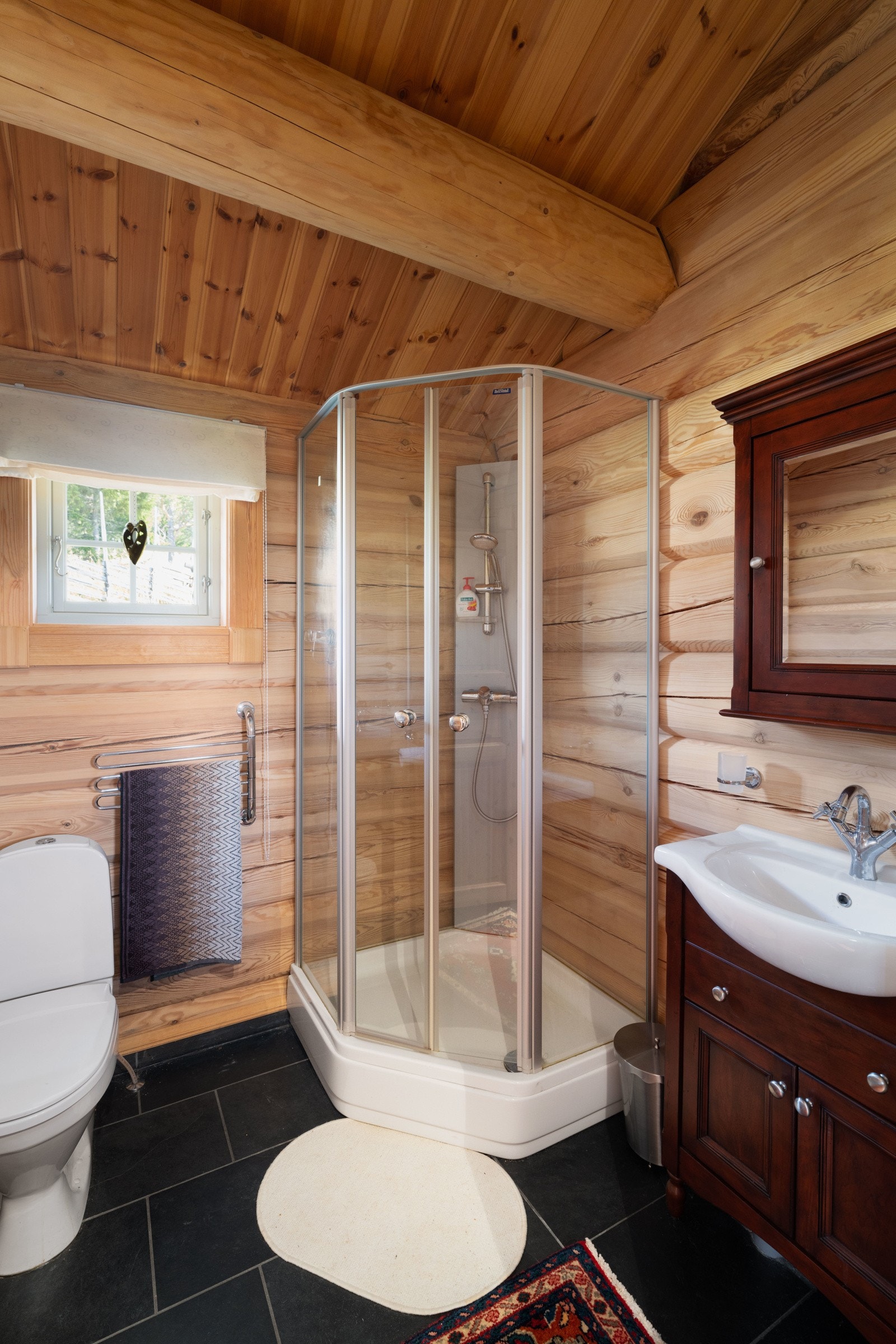Annekset har også et eget dusjbad med gulvstående vannklosett, dusjkabinett, servant med over- og underskap i malt tre. Sorte moderne fliser på gulvet med varmekabler i gulv. Tømmervegger og mønet himling. Galleribilde