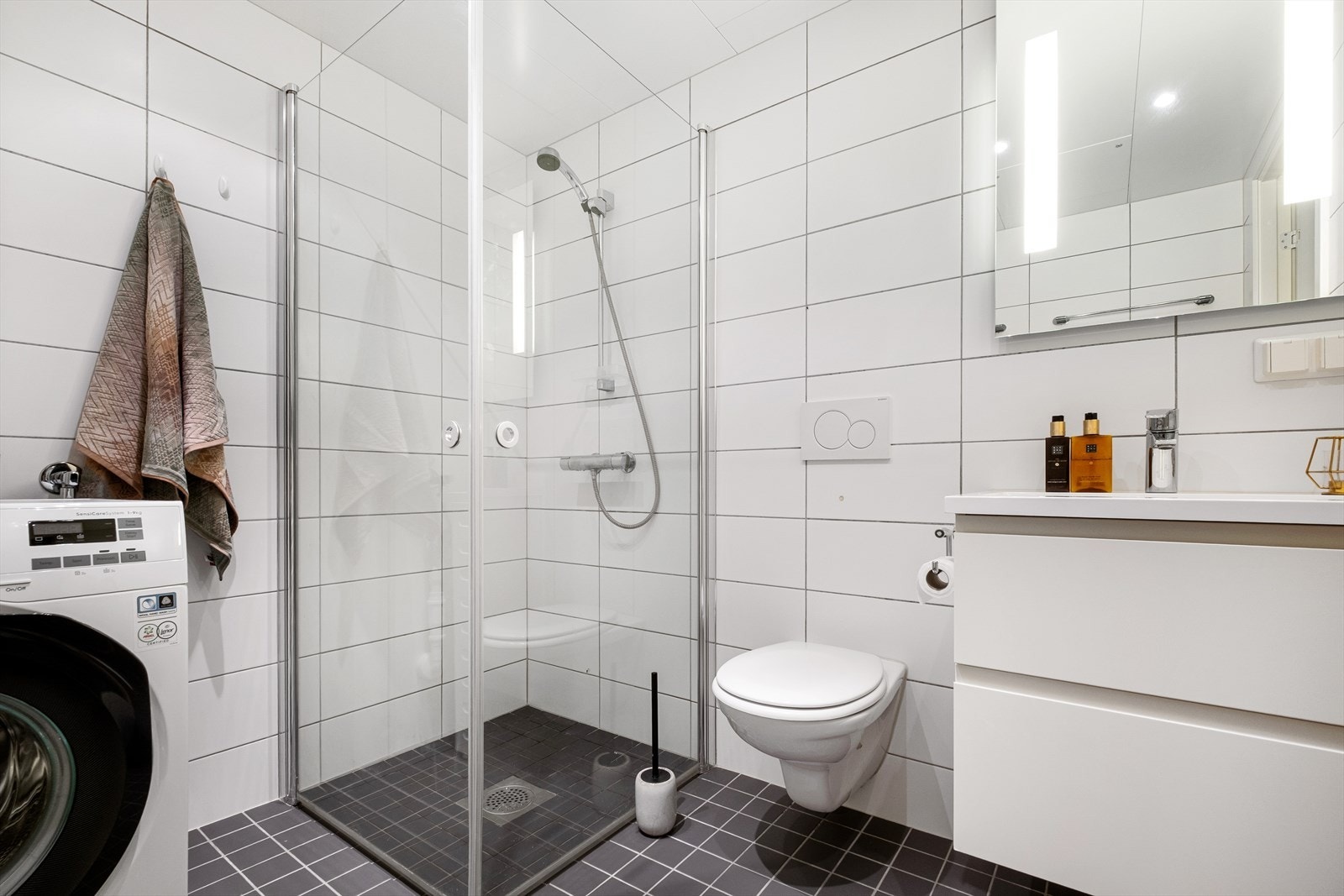 Badet er innredet med vegghengt toalett, servant med innredning, speil med belysning og dusjhjørne med svingbare glassdører. Galleribilde