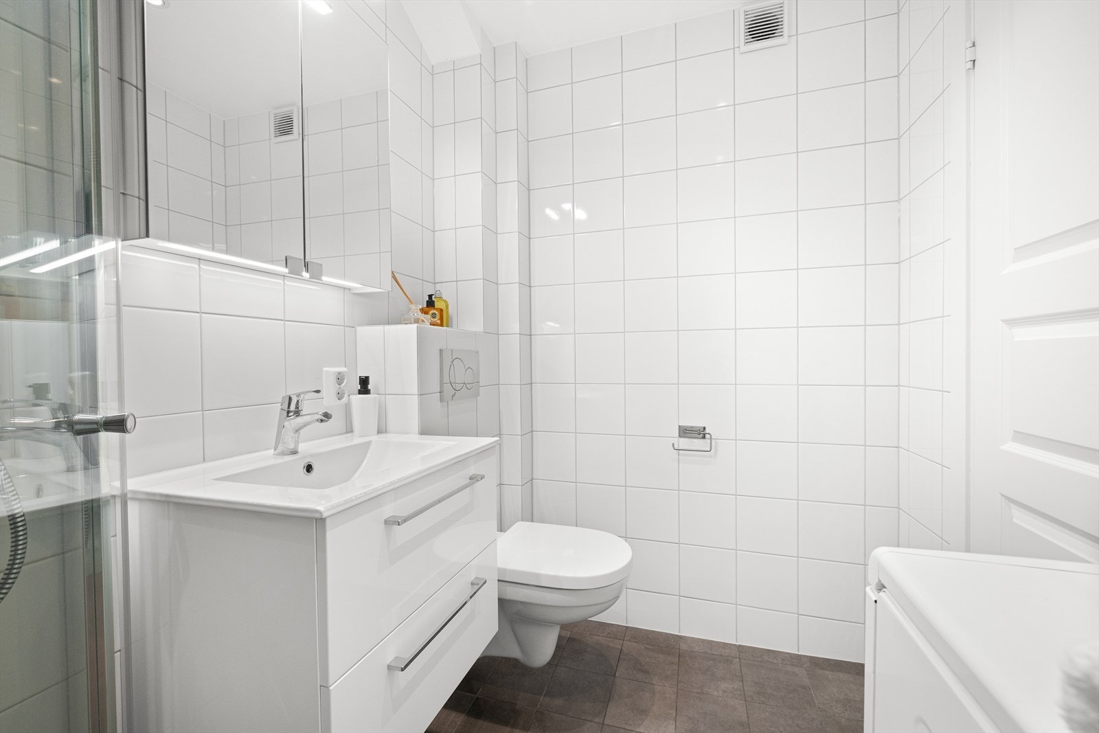 Innredet med vegghengt toalett og innebygget sisterne, samt stilren servant med hvite høyglans fronter, porselenservant, ettgreps armatur og speilskap med integrert belysning. Det er opplegg og plass til vaskemaskin. Galleribilde