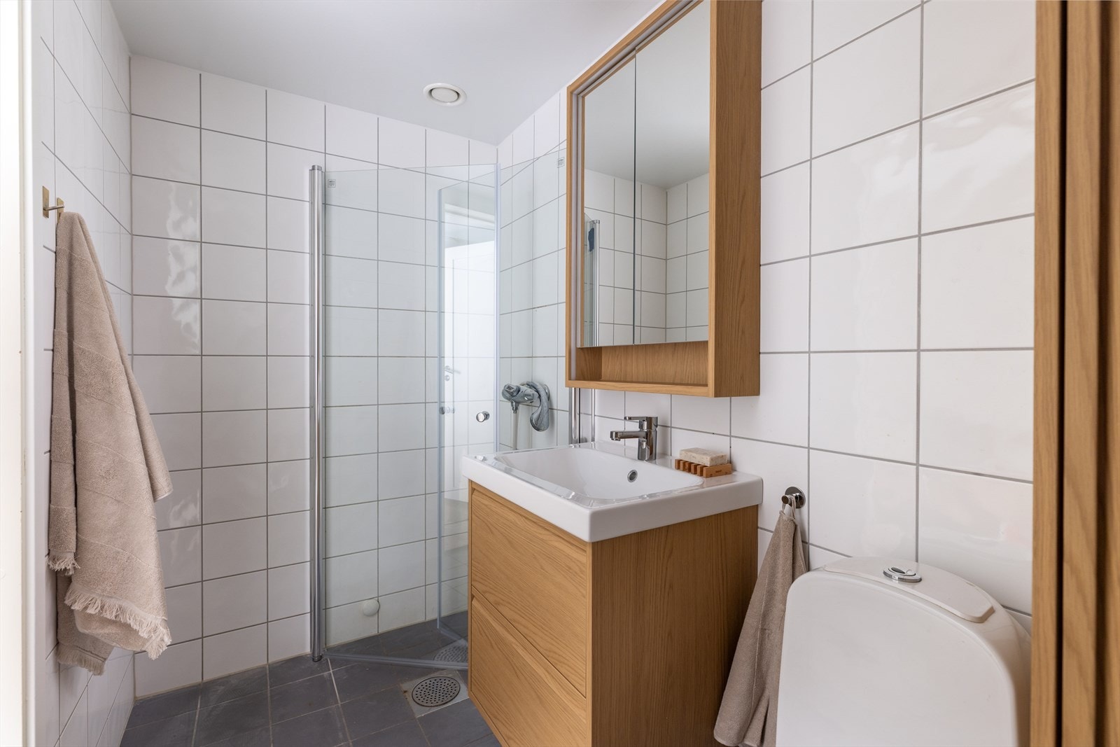 Badet er utstyrt med toalett, servant, overhengt skap med speil, dusjsone med innfellbare glassdører, og mekanisk avtrekk Galleribilde
