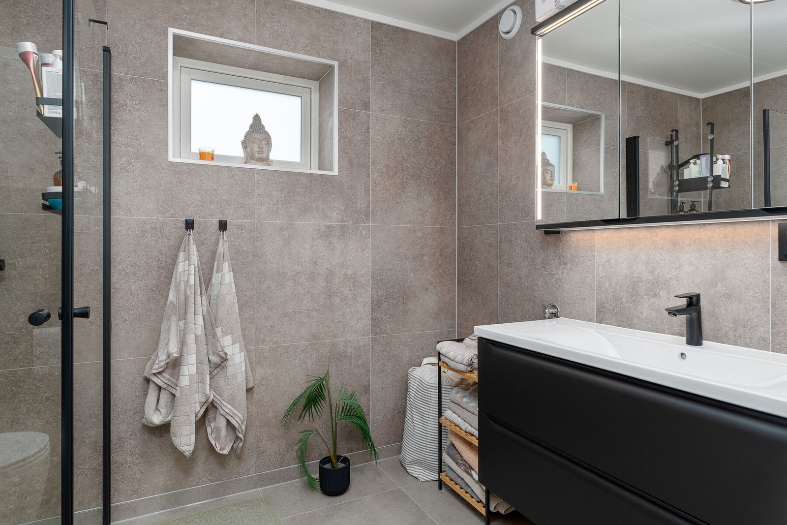 Moderne bad med servant på skuffeseksjon, speilskap med lys, vegghengt toalett og dusjhjørne. Det er opplegg for vaskemaskin. Galleribilde