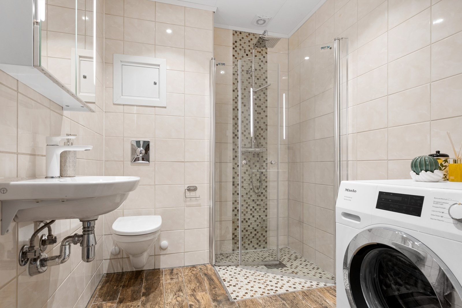 Pent bad med flislagte gulv og vegger, samt downlights i taket. Badet er utstyrt med servant, speilskap, vegghengt toalett og dusjhjørne med innfellbare glassdører. Galleribilde