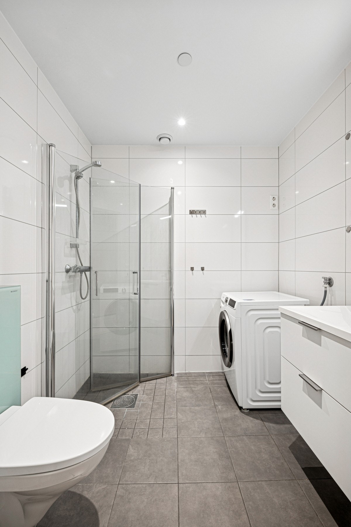 Funksjonelt bad med dusjhjørne med glassdører, vegghengt toalett og opplegg for vaskemaskin. Praktisk og gjennomtenkt løsning i moderne utførelse. Galleribilde