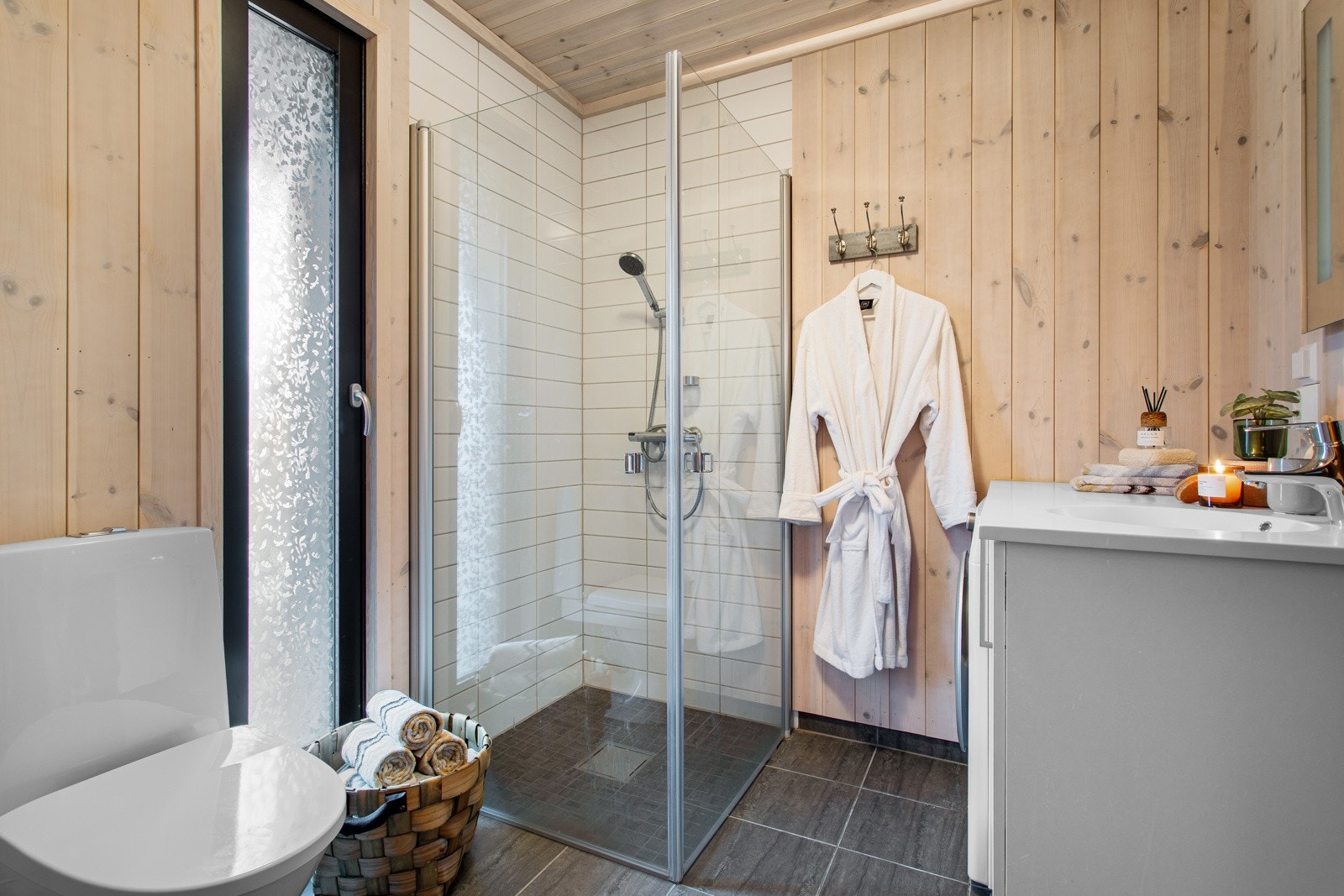 Lyst og nordisk bad med flis på gulv og panel på vegger. Badet inneholder innredning med nedfelt servant, dusjnisje med glassvegger og toalett Galleribilde