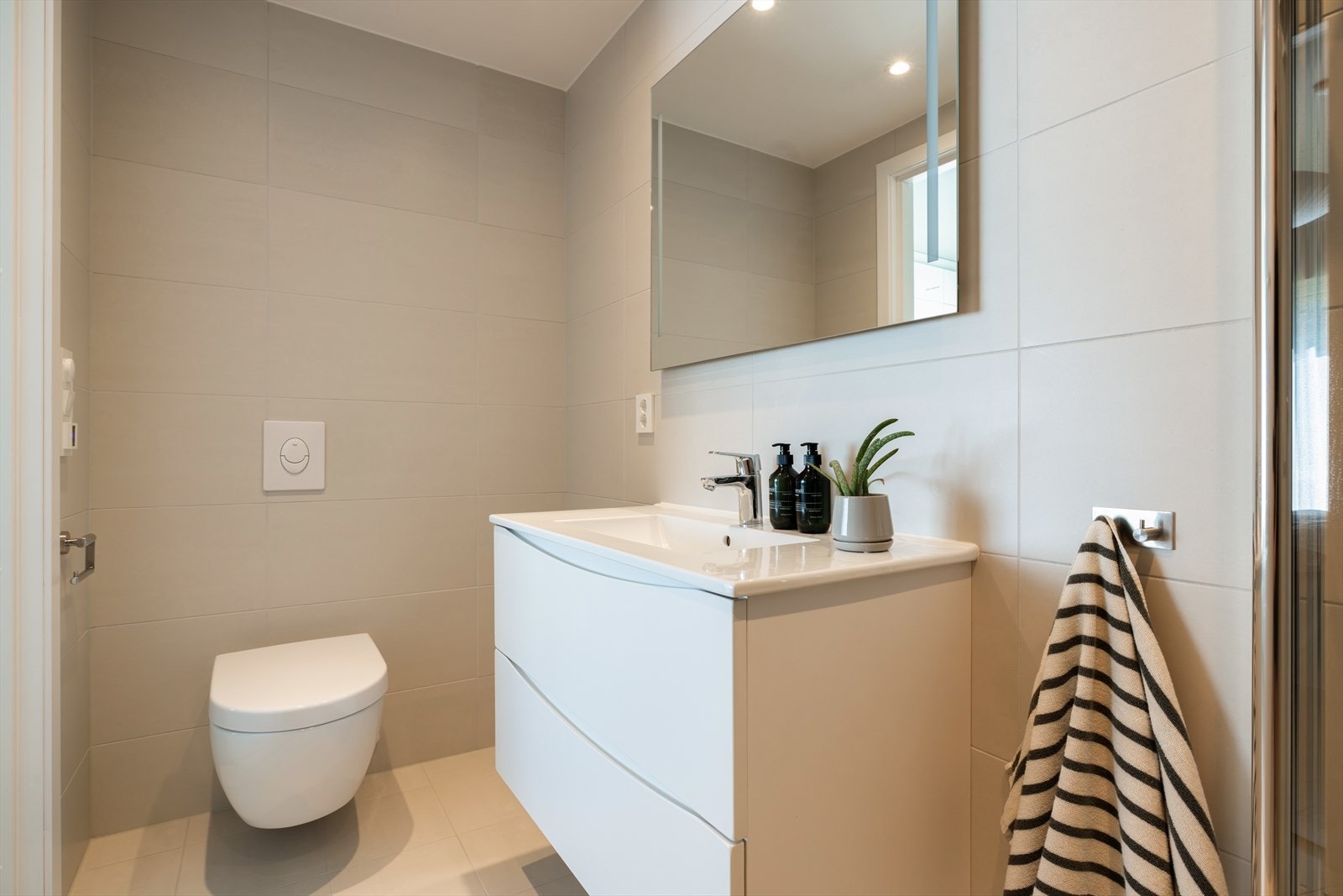 Innredningen består av en heldekkende servant på hvit skuffeseksjon, speil med integrert belysning og et veggmontert toalett. Galleribilde