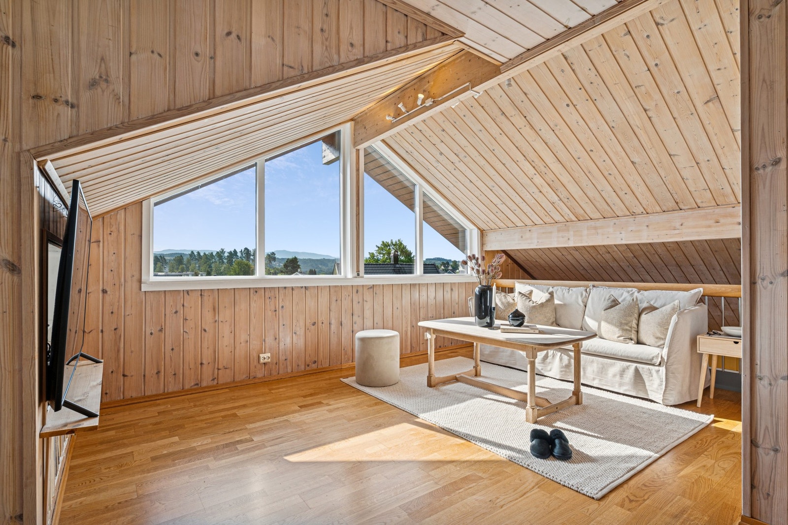 Lys og lun stue med skrå himling og store vindusflater som slipper inn rikelig med dagslys og gir utsikt mot åsene i horisonten. Galleribilde