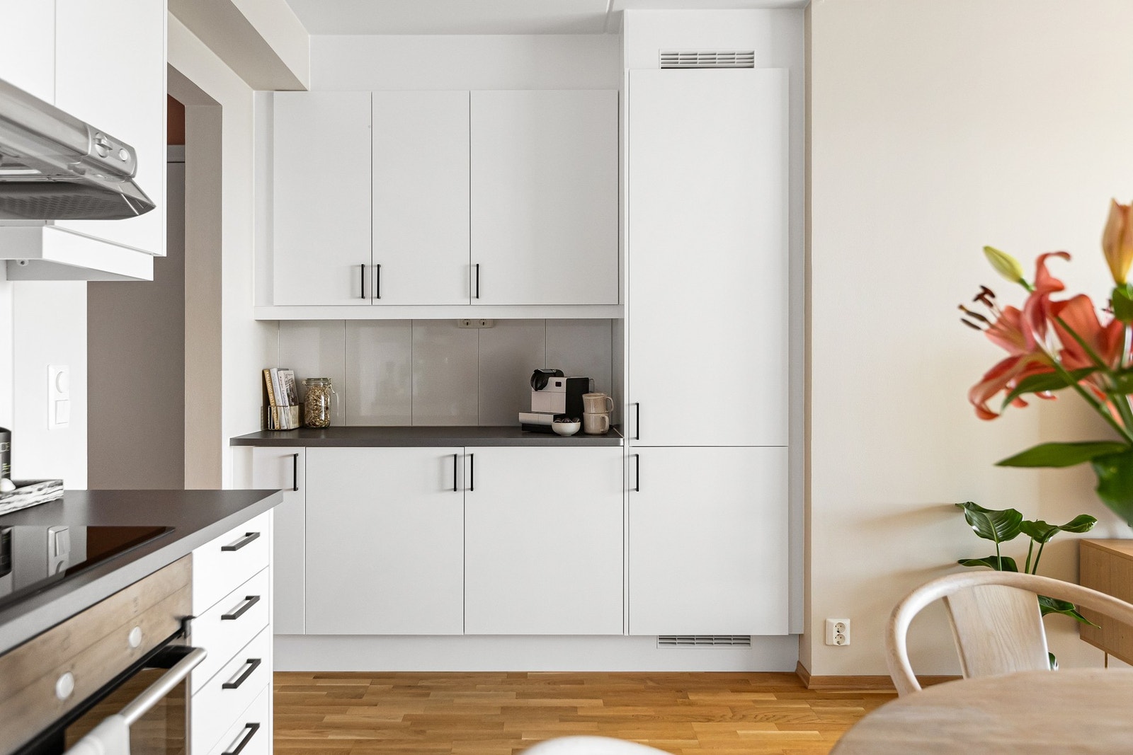 Kjøkkeninnredningen har hvite glatte fronter, laminat benkeplate med planlimt kum i rustfritt stål, flisfelt på vegg over kjøkkenbenk og belysning under overskap. Galleribilde